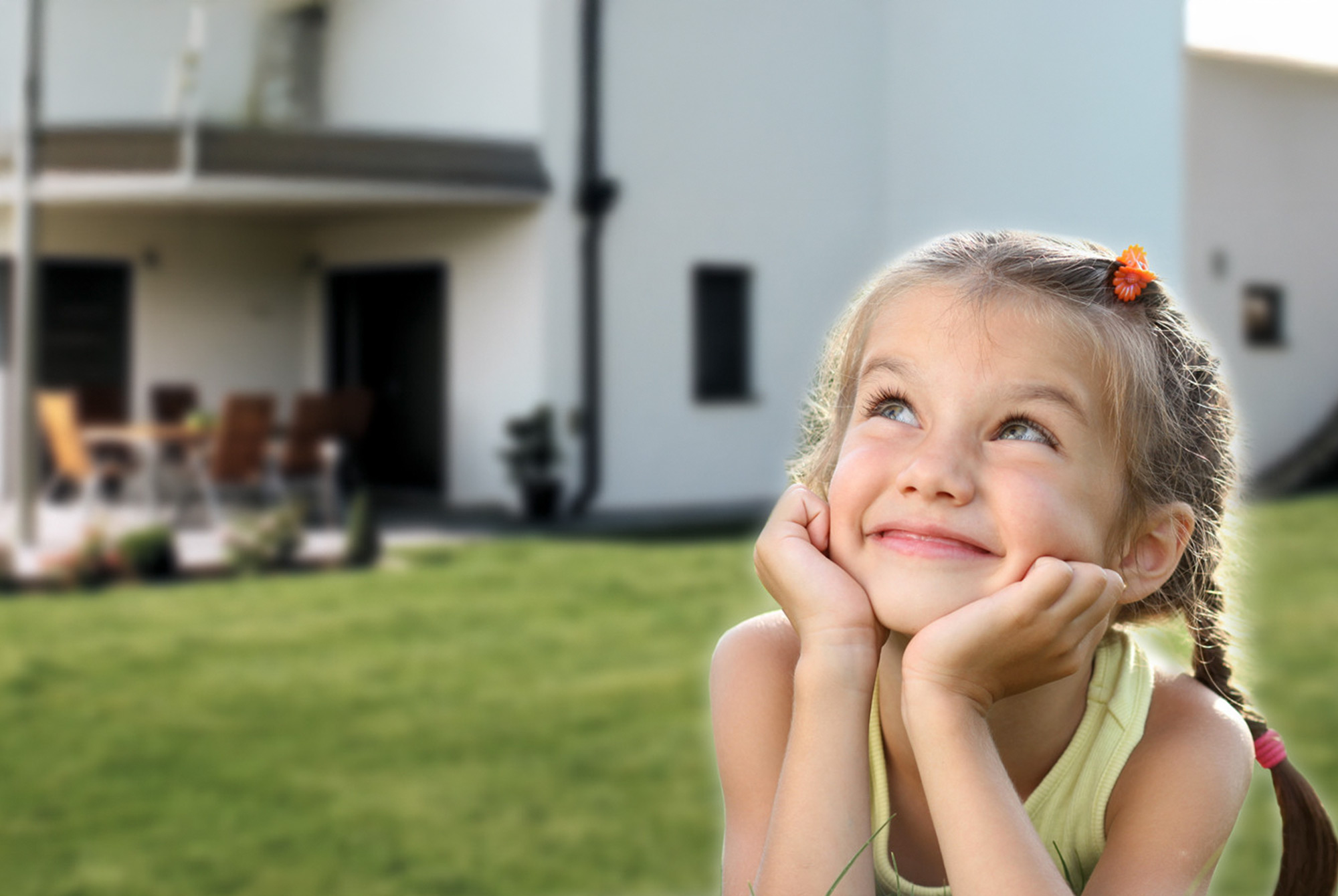 Fröhliches Mädchen liegt auf dem Rasen vor einem Einfamilienhaus und blickt lächelnd nach oben.
