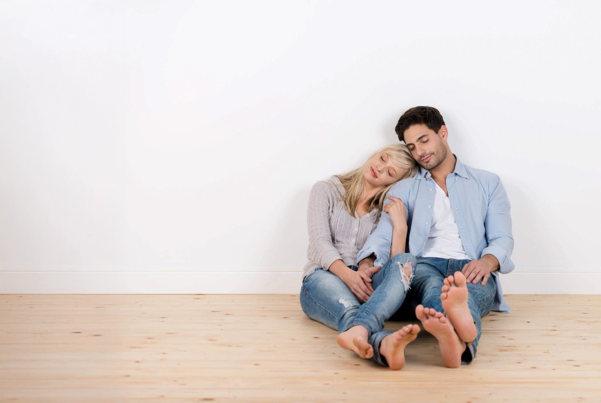 Junges Paar sitzt barfuß und entspannt an einer weißen Wand in einem leeren Zimmer auf Holzboden.